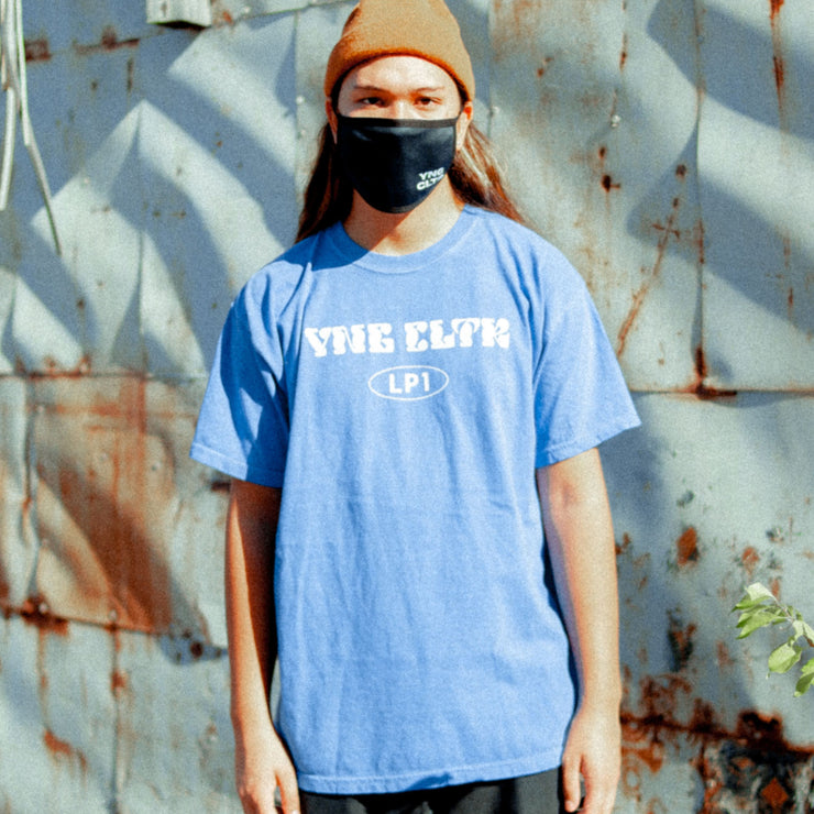 Periwinkle short sleeve shirt with "YNG CLTR" written across the chest in white bubble lettering. Below that is an oval, and inside "LP1" is written. An individual is wearing the short sleeve shirt and standing in front of a wall.