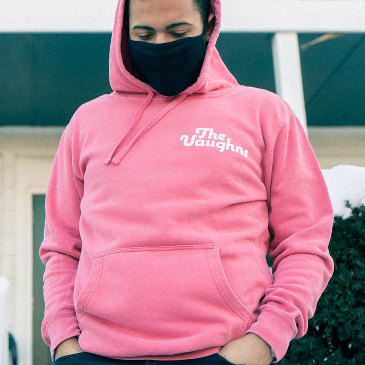 Salmon colored pullover hoodie with white text in the top corner of the chest that says THE VAUGHNS. Down one of the sleeves there is white text that says ROM-COMS & TAKE-OUT. An individual is modeling the hoodie and standing in front of the front porch to a house.