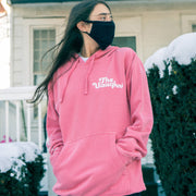 Salmon colored pullover hoodie with white text in the top corner of the chest that says THE VAUGHNS. Down one of the sleeves there is white text that says ROM-COMS & TAKE-OUT. An individual is modeling the hoodie and standing in front of the front porch to a house.