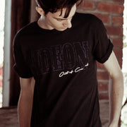 Black short sleeve shirt with light purple text across the chest that says GIDEON. Overlapping the purple text on the bottom right is white cursive text. An individual is modeling the shirt and standing in front of a brick wall.