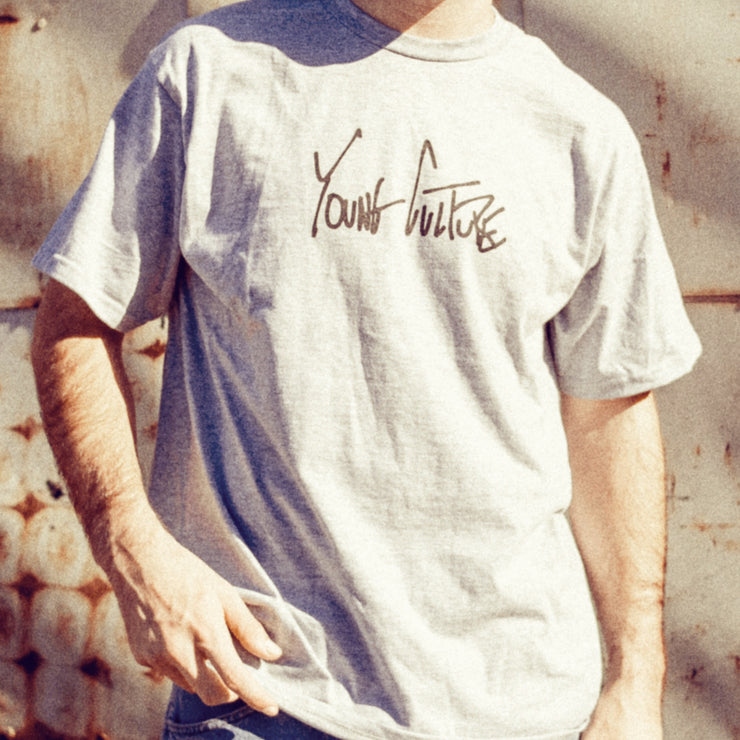 Athletic grey short sleeve shirt with YOUNG CULTURE written across the chest in black lettering. An individual is modeling the shirt and stranding in front of a concrete wall.