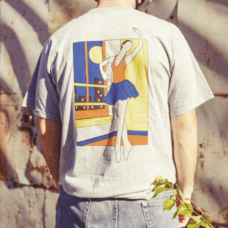 Back of the short sleeve shirt is a drawing of a ballerina in red blue and yellow. She is dancing in a room, and outside the window are tall buildings and the moon in the sky. An individual is modeling the shirt and standing in front of a concrete wall.