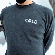 Male Model wearing a Dark charcoal grey pullover crewneck. The word COLD is embroidered in white thread on the top right part of the crewneck. In the center of the O in Cold are 5 nails, Gideon's symbol.