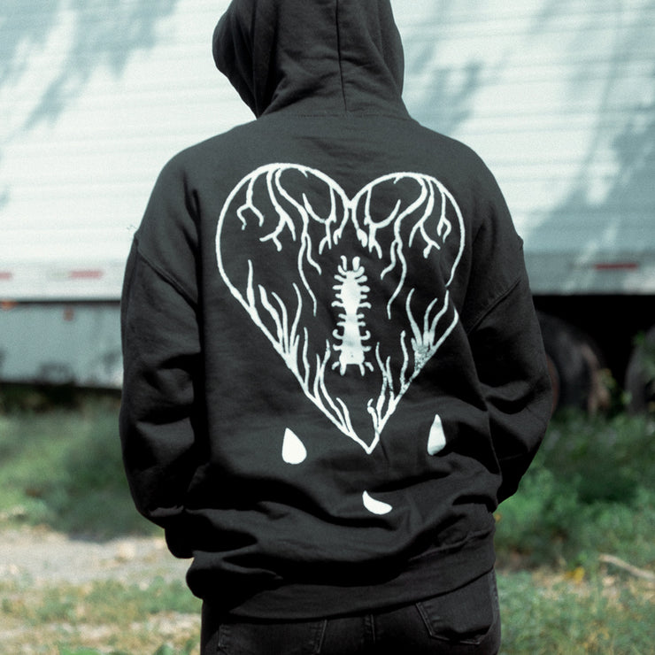 Back of black pullover hoodie. There is a large white heart with veiny lines and an insect inside. Below that is three droplets falling. There is a model wearing the hoodie with their back turned away from the camera and standing in front of a truck.