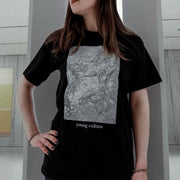 Black short sleeve shirt with a rectangle design filled with white and grey marble pattern. Below that there is small white text that says young culture. An individual is modeling the shirt and standing inside against a white pole.