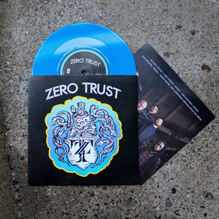 Black vinyl jacket with ZERO TRUST written across the top. Below the text is a drawing of a skeleton with snakes surrounding it, drawn in white. Below the skeleton is a crest that has the letters Z and T written inside it. Peeking out of the top of the vinyl jacket is a blue vinyl. Both are laying on an asphalt surface.