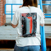 Back of cream colored short sleeve shirt with text along the side that says CALLING ALL CAPTAINS. In the center there is artwork of a coffin laying in grass surrounded by trash. Below that there is text that says SLOWLY GETTING BETTER. An individual is modeling the shirt and leaning against a railing on top of stairs, with a brick building to the side.