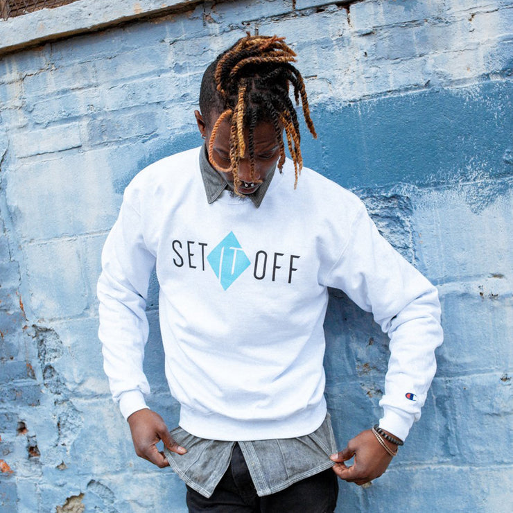 White crewneck with text across the chest that says SET IT OFF. The word IT is written inside of a blue diamond and in white text, and the words SET and OFF are written in black text. An individual is modeling the crewneck and standing in front of a blue concrete brick wall.