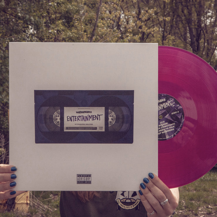 White vinyl jacket with a purple cassette tape in the center. The cassette tape has purple text on it that says ENTERTAINMENT. Peeking out of the jacket is a transparent purple vinyl. An individual is holding both, and standing in front of trees and a forest.