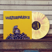 Vinyl jacket with a picture a blue grenade on it and a yellow background.  Text on the cover reads Waterparks. Sticking out of the vinyl cover is a yellow mix colored record. The record is leaning on the side of a house on what is a back porch.