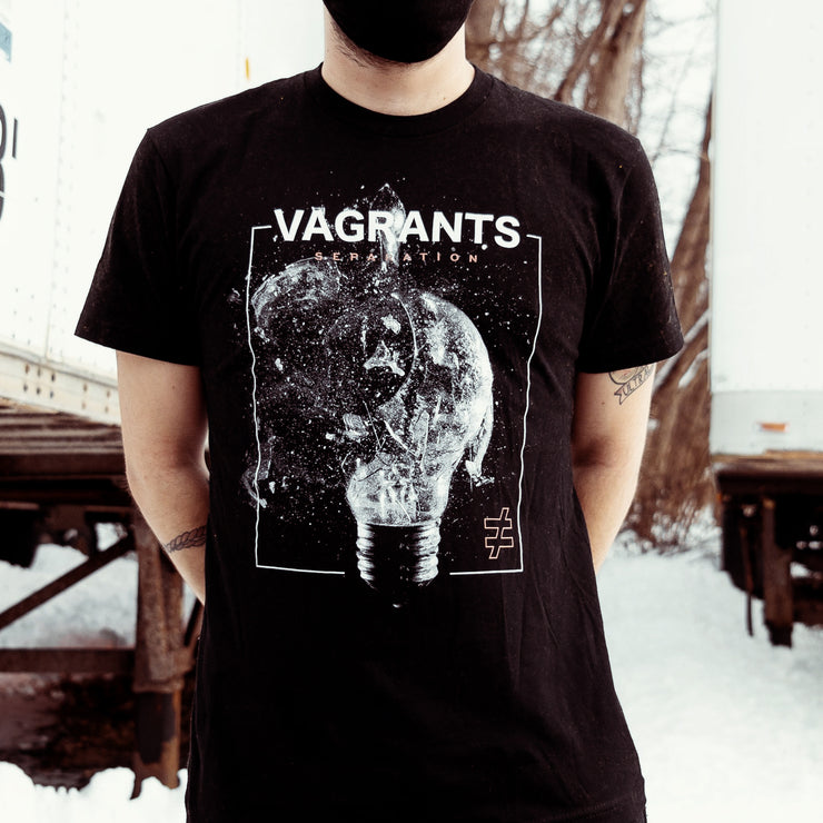 Black short sleeve shirt with a box drawn in the center using white lines. Inside the box, there is a black and white lightbulb being shattered. In the bottom right there is a not equal to sign drawn in color. On the top of the box, it says "VAGRANTS" in large white font. The shirt is being worn by a model standing outside between two trucks, with snow on the ground.