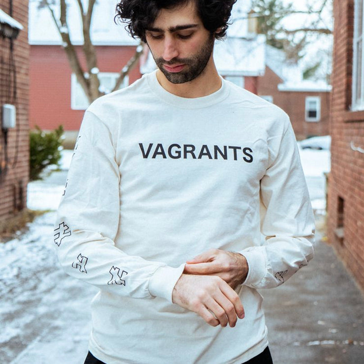 Cream colored long sleeve shirt with VAGRANTS written across the chest in black font. Along the sleeves are black outlined not equal symbols. An individual is modeling the shirt and standing in between two brick buildings with snow on the ground.