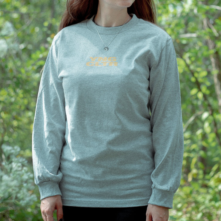 Grey long sleeve shirt with YNG CLTR in orange on the chest. On the back there is a ca drawn in orange with a triangle around it. Above that, there is text that says GODSPEED, also printed in orange. An individual is modeling the shirt, standing in front of greenery.