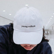 Plain White Dad Hat with "young culture" written on front with in black lowercase lettering. Hat is being modeled by an individual with dark brown hair in front of a white background.