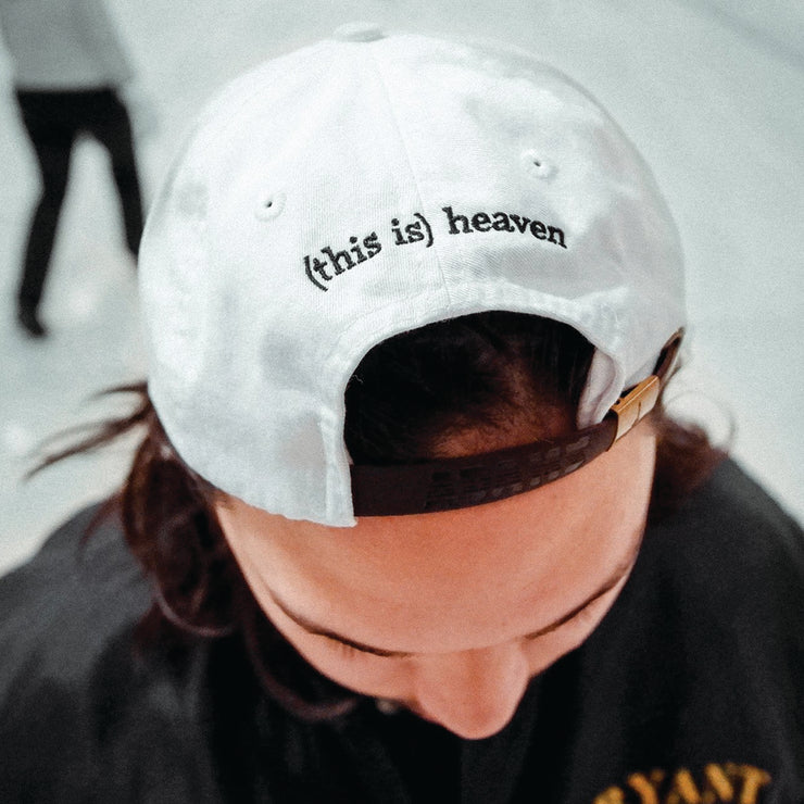 Back of a Plain White Dad Hat with lowercase black text writing "(this is) heaven". Hat is being modeled by an individual with dark brown hair in front of a white background.