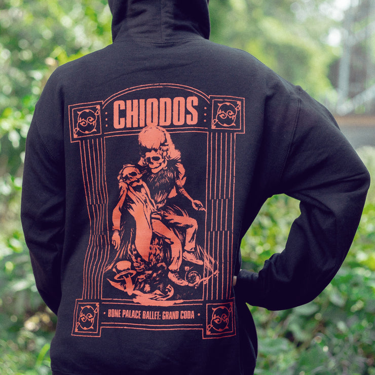 Back of black pullover hoodie with a drawing of two skeletons, one holding the other in red. On top of the skeletons there is red font that says CHIODOS. An individual is modeling the hoodie and standing in front of trees and leaves.