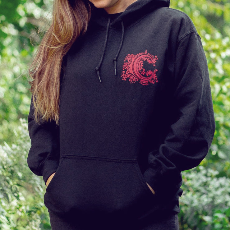 Black pullover hoodie with an embellished letter C in the top corner drawn in red. An individual is modeling the hoodie and standing in front of trees and leaves.