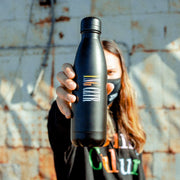 Black stainless steel water bottle with partially colored lettering spelling "YNG CLTR". "YNG" is colored yellow, pink, and orange, and CLTR is colored white. Water bottle is being held by an individual wearing a black face mask and a Young Culture shirt.