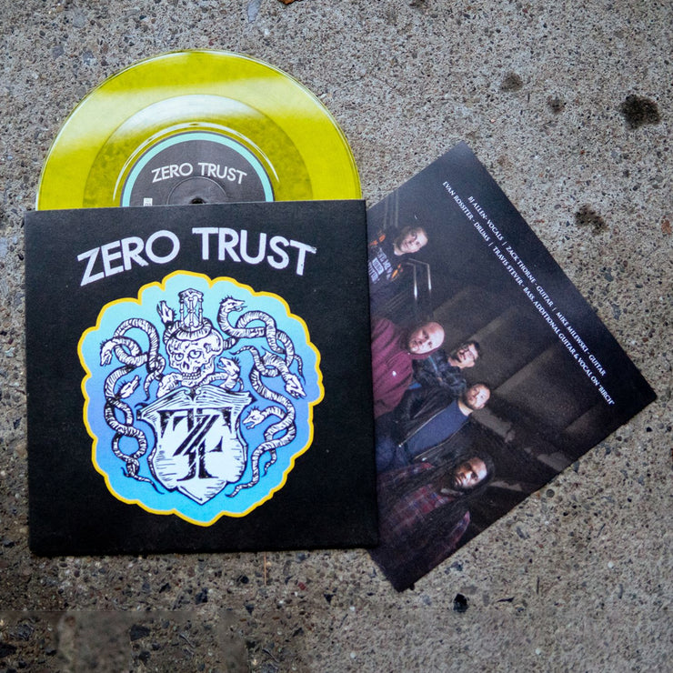 Black vinyl jacket with ZERO TRUST written across the top. Below the text is a drawing of a skeleton with snakes surrounding it, drawn in white. Below the skeleton is a crest that has the letters Z and T written inside it. Peeking out of the top of the vinyl jacket is a yellow vinyl. Both are laying on an asphalt surface.